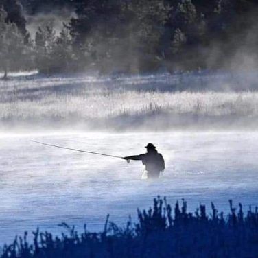 Silhouette of a person fly fishing in a misty river at dawn