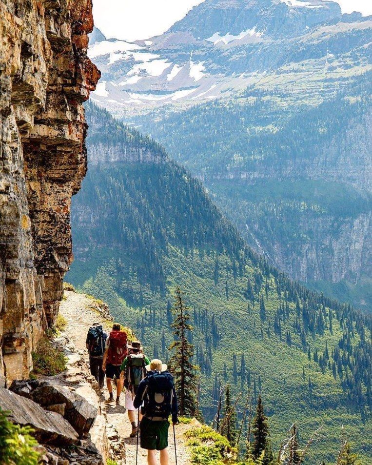Hikers on a narrow mountain trail beside a cliff, overlooking a green valley and distant peaks.