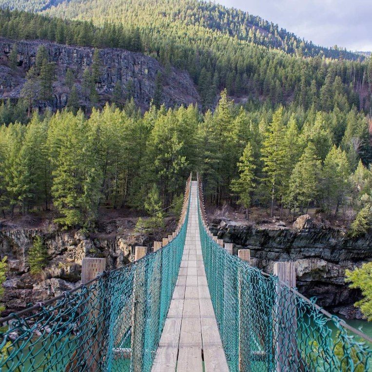 Suspension bridge with turquoise railings over a rocky gorge in a forested mountain valley