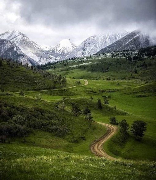 Winding dirt path through green hills toward snow-covered mountains under cloudy sky