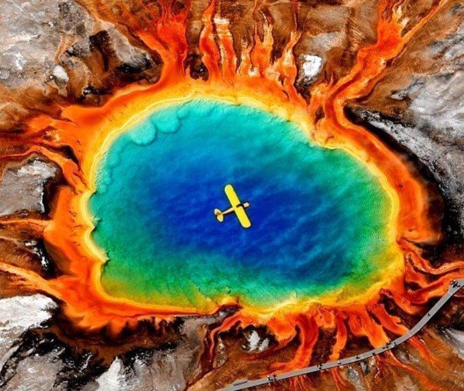 Aerial view of a blue-green hot spring with orange microbial mats and a yellow plane overhead