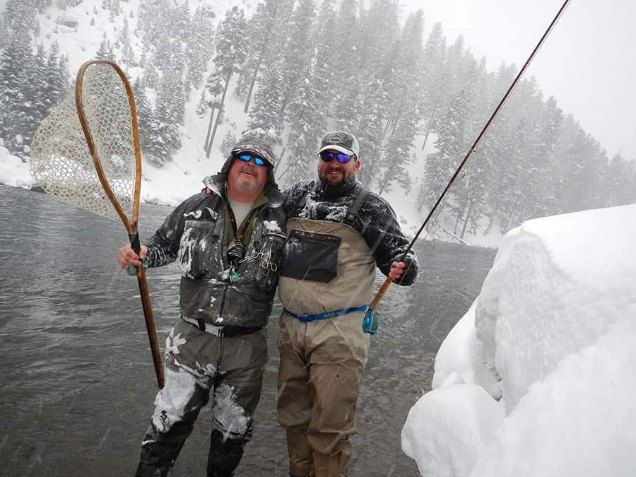 Two people fishing in a snowy river beside a large snowbank, one holding a landing net and the other a rod.