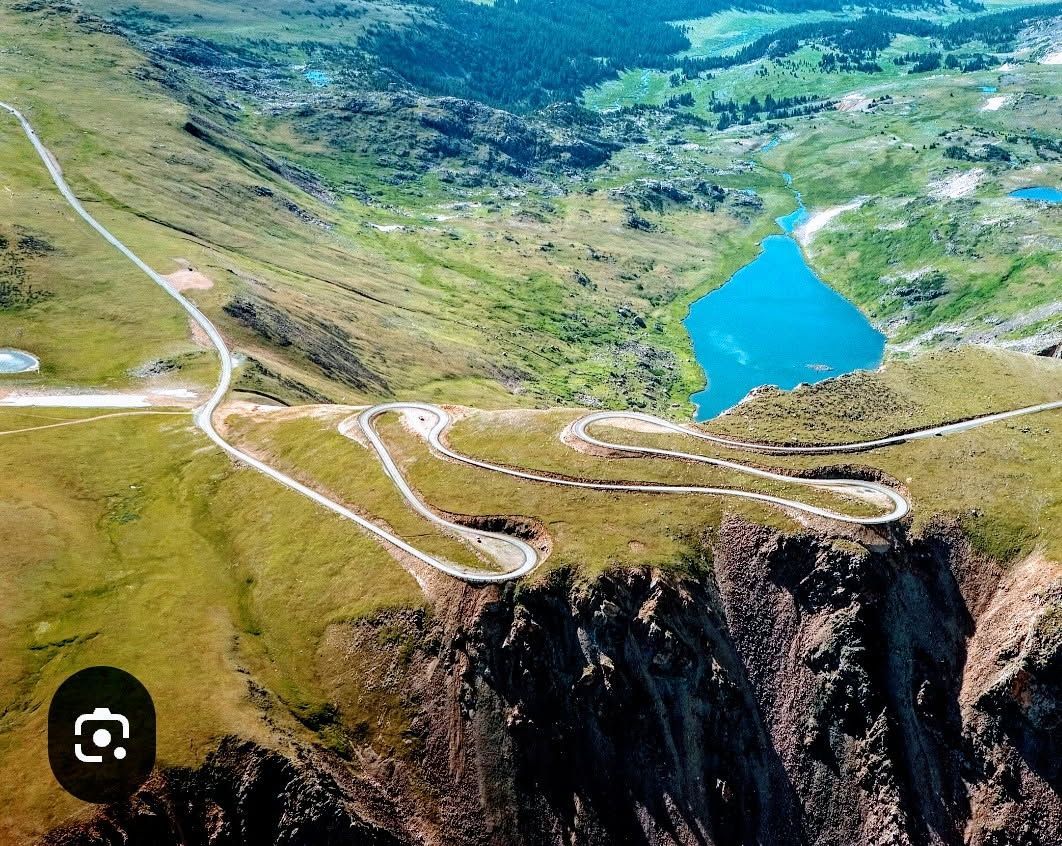 Aerial view of a winding mountain road beside a bright blue lake and grassy cliffs