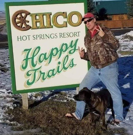 Man posing by Chico Hot Springs Resort sign reading “Happy Trails,” with a dog at his feet in snowy daylight