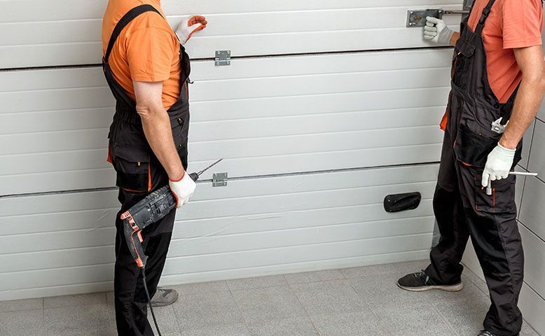 Two workers in orange shirts and black overalls standing by a white garage door, holding tools.