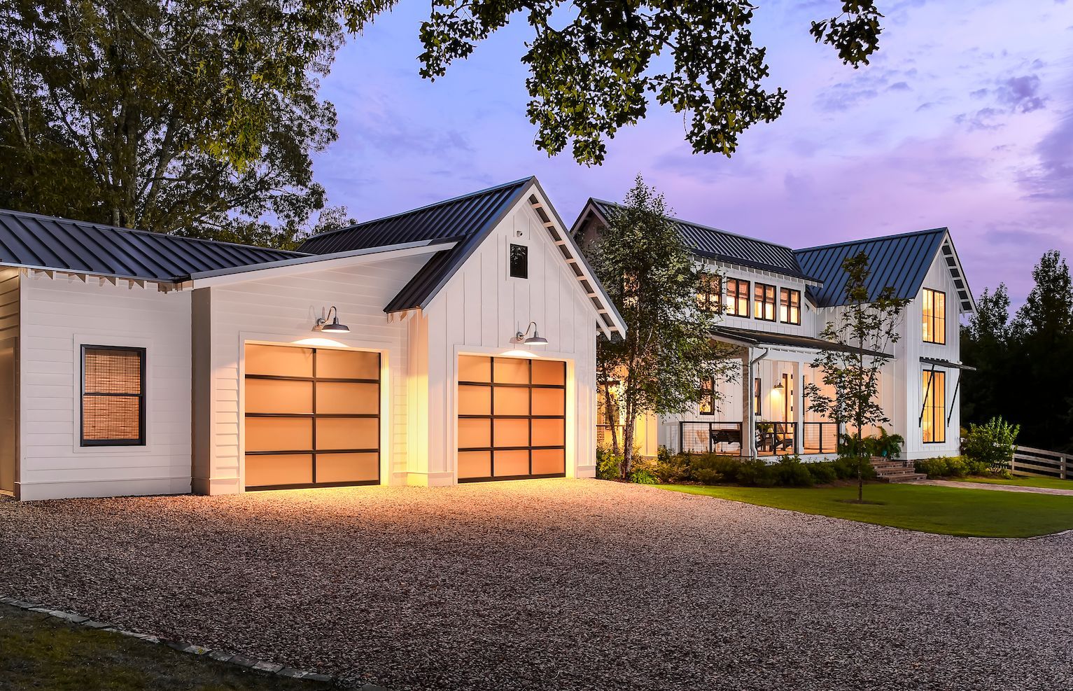 Modern white house with lit garage doors, gravel driveway, and trees at dusk