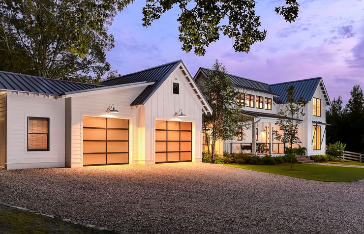 Modern white house with lit garage doors, gravel driveway, and trees at dusk