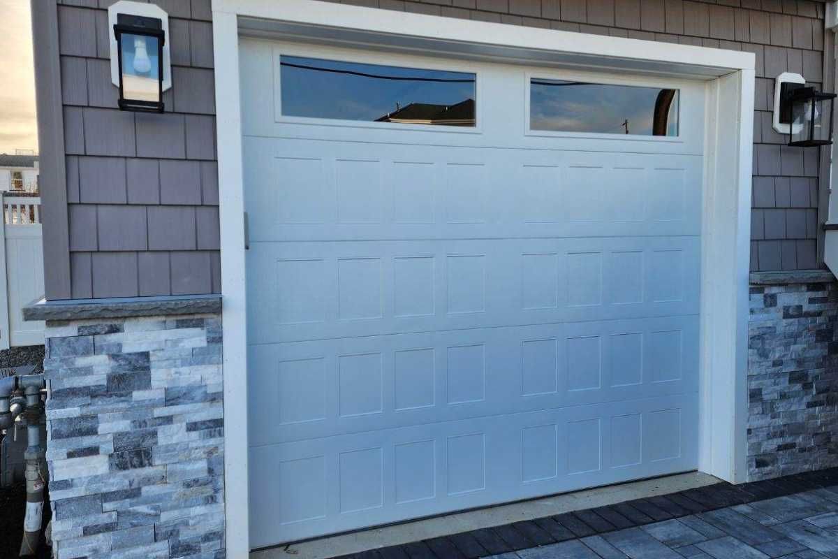 White garage door with small windows on a house exterior, framed by gray siding and stone trim