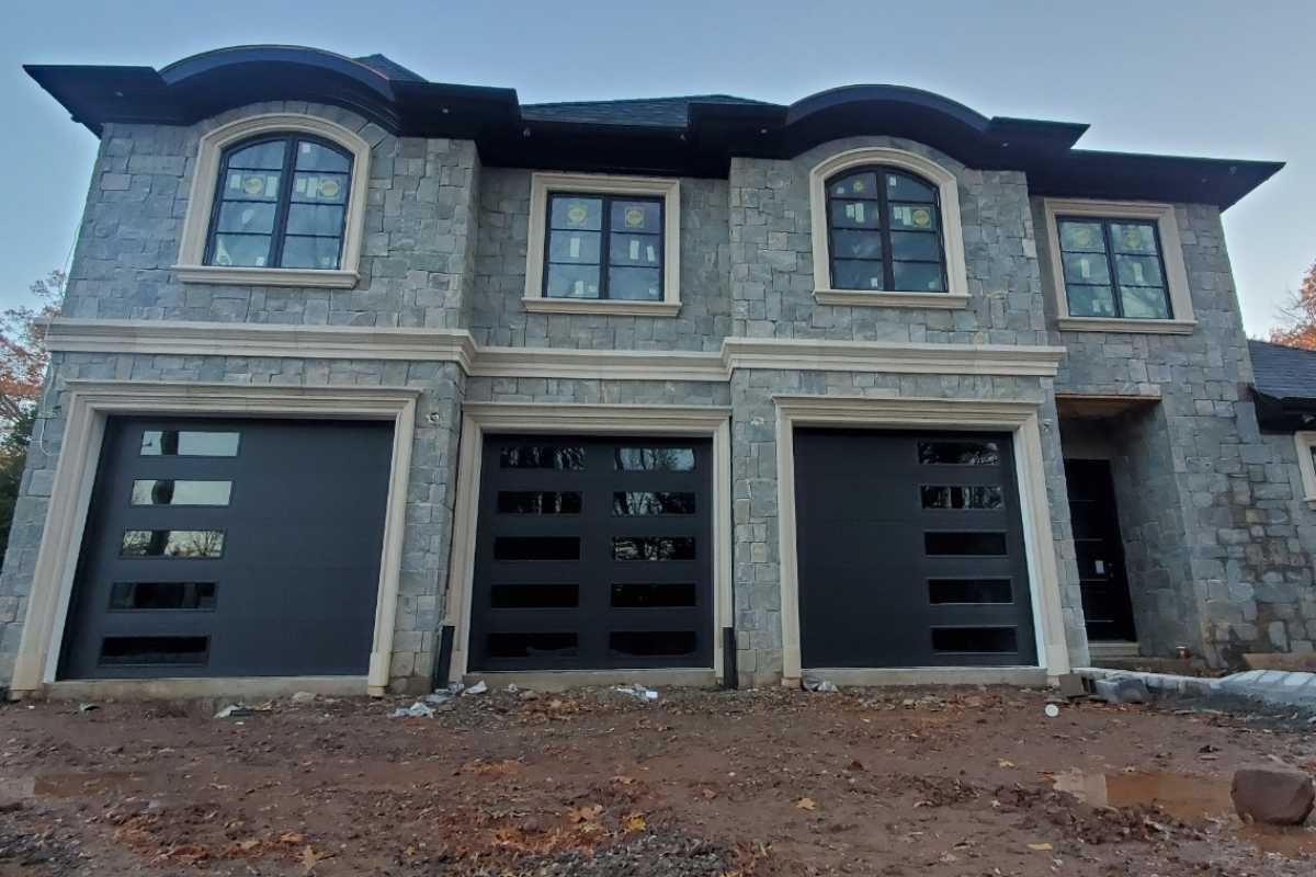 Stone house exterior with three black garage doors and arched upper windows, unfinished front yard