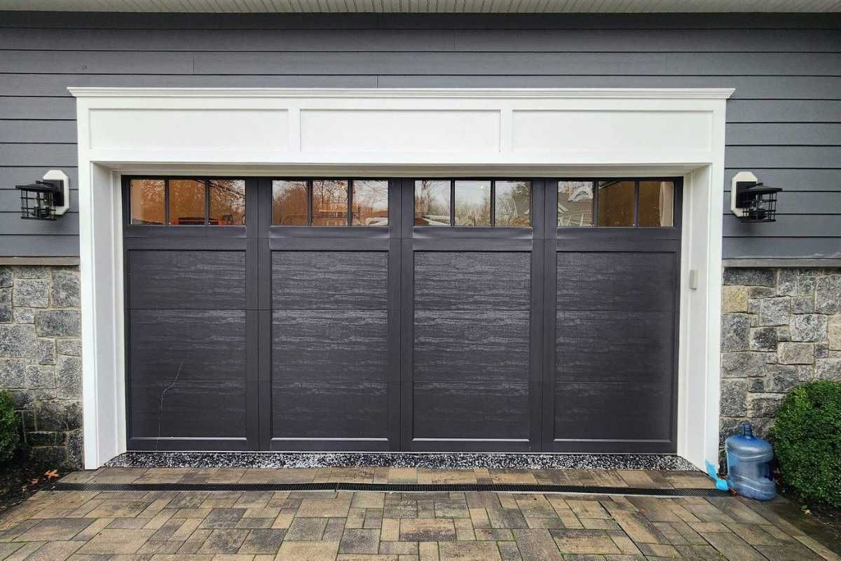 Black garage door with white trim on a gray stone-front house, with brick pavers in front