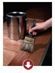 A paintbrush being dipped into a metal paint can on a wooden floor, with a red down arrow icon below.