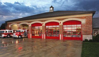 Fire station with red bay doors and fire trucks parked outside on a wet pavement at dusk