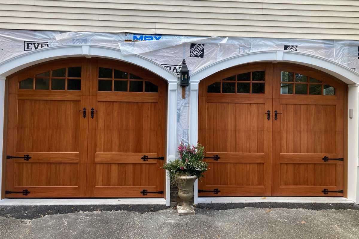 Two wooden garage doors with arched tops beneath a white stucco facade and central potted flowers