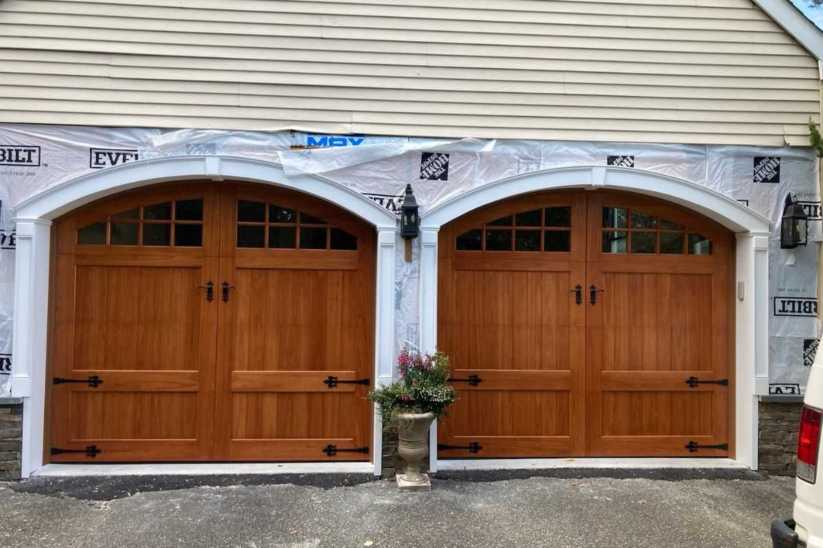Two wooden garage doors on a house exterior with white trim and a small potted plant between them
