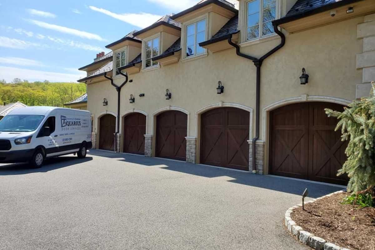 Row of beige townhouses with wooden garage doors, beside a parked white van on a sunny street.