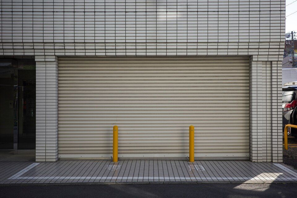 Closed beige roll-up garage door in a tiled building facade, with two yellow bollards in front.