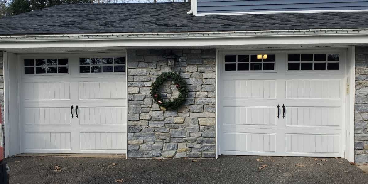 Two white garage doors with stone center panel and wreath on a gray house facade