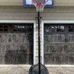 Basketball hoop in front of two closed gray garage doors, with a driveway and black pole base.