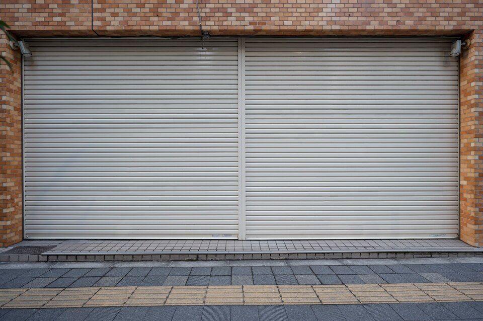 Closed gray metal storefront shutters on a brick building above a tiled sidewalk