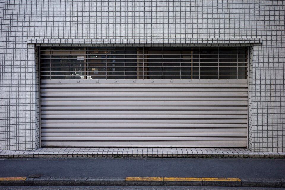 Closed gray storefront shutter with a narrow barred window above it along a street curb