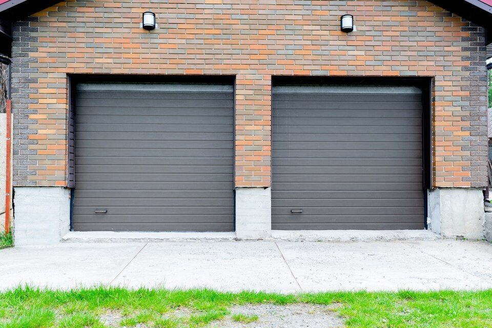 Two gray garage doors in a brick building with wall lights and a concrete driveway
