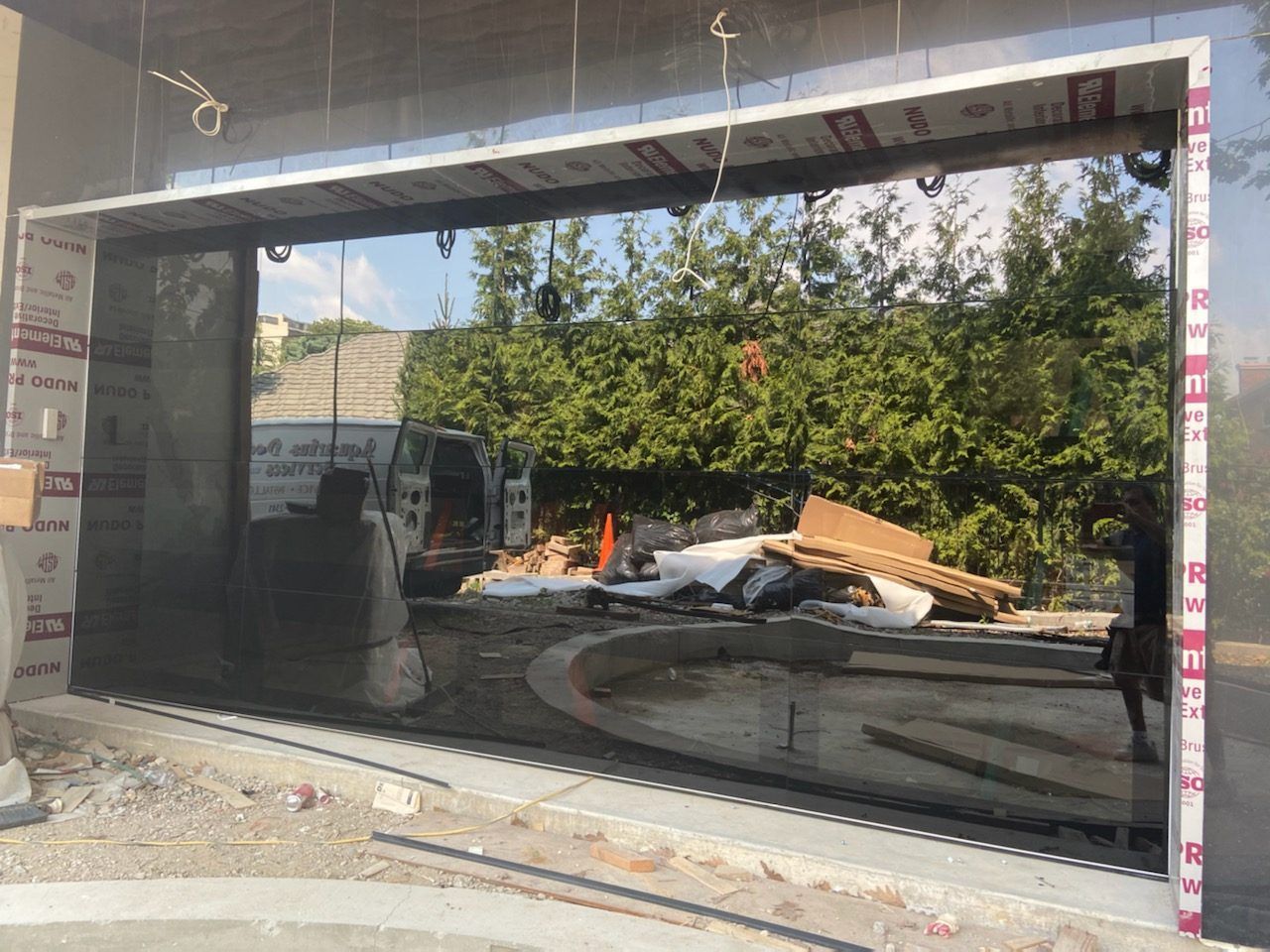 Construction site with a large glass storefront frame and scattered debris beside trees.