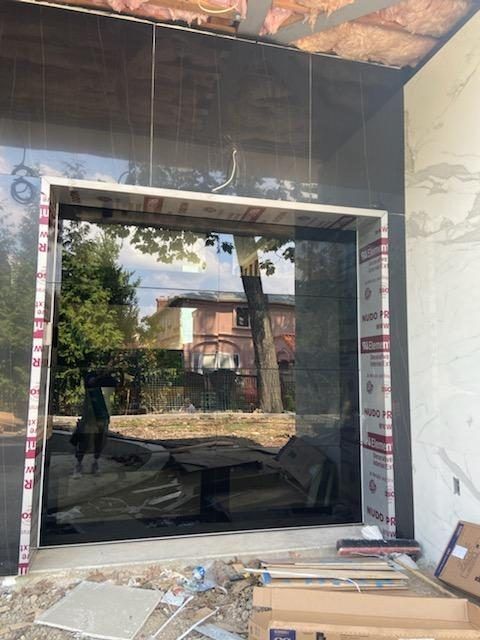 Large black-framed glass window installation at a construction site, reflecting trees and a building outside.