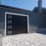 Modern house exterior with open garage door, stone wall, and paved driveway.