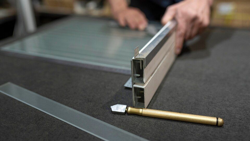 Hands positioning a metal paper cutter beside a glass pane and utility knife on a workbench