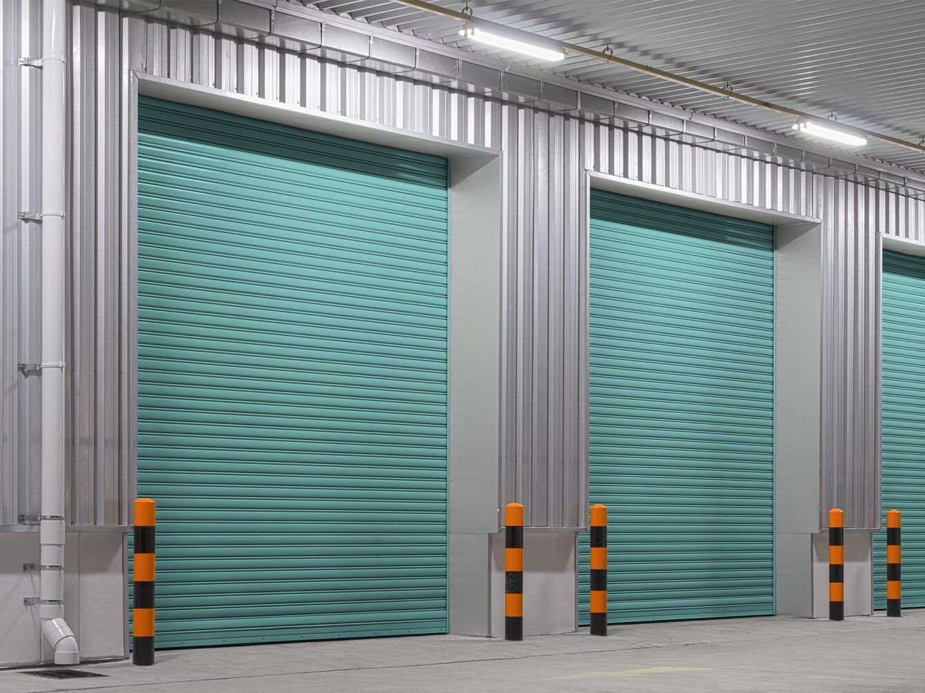 Teal roll-up industrial doors in a warehouse with metal siding and orange safety bollards