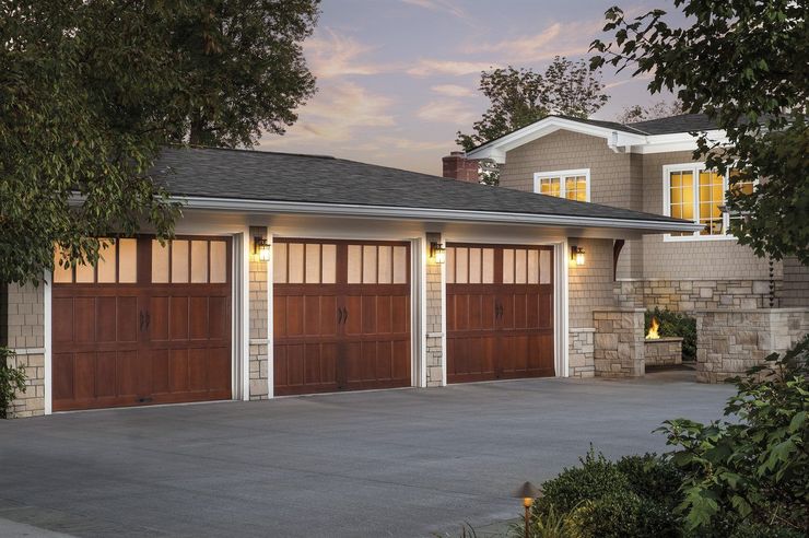 Three-car garage with wooden doors and stone exterior at dusk, lit driveway and home entrance