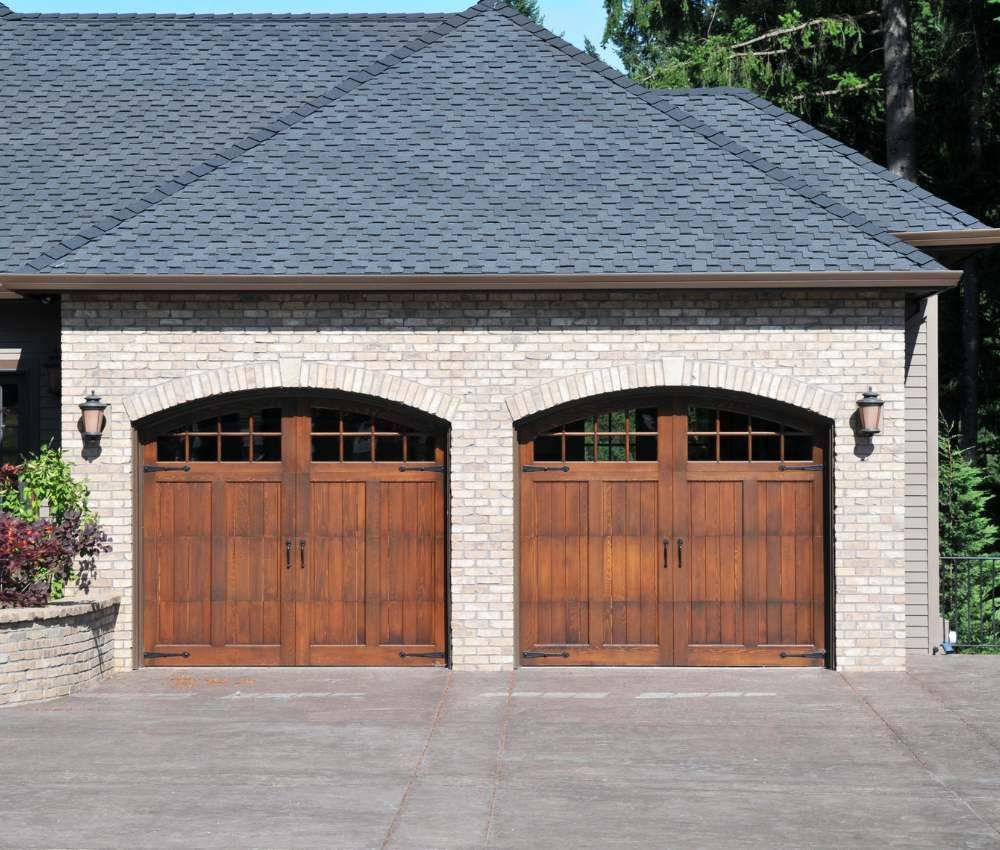 Two wooden garage doors on a brick house with a gray shingled roof and paved driveway