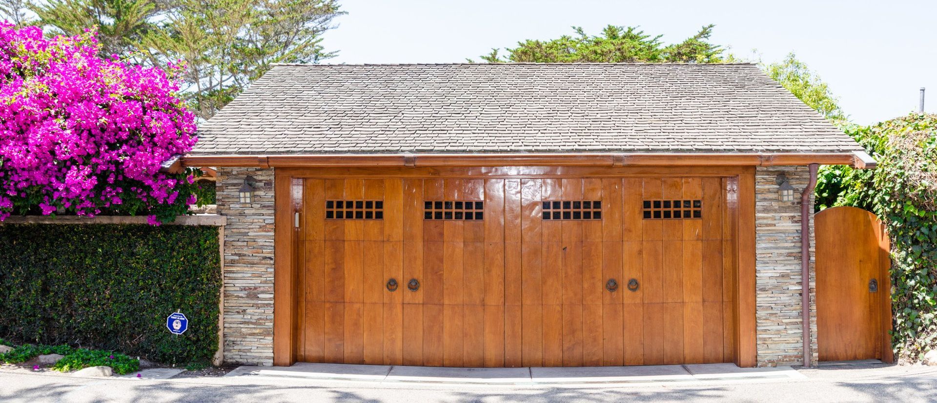 Wooden garage doors beneath a tiled roof, framed by pink bougainvillea and hedges on a sunny street