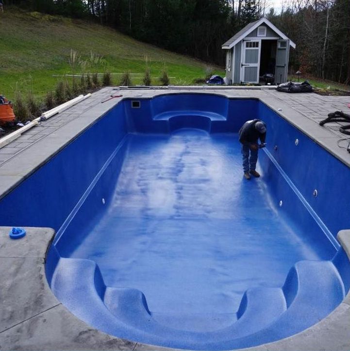 Person working inside a bright blue swimming pool. Gray concrete border surrounds pool. Small shed in background.