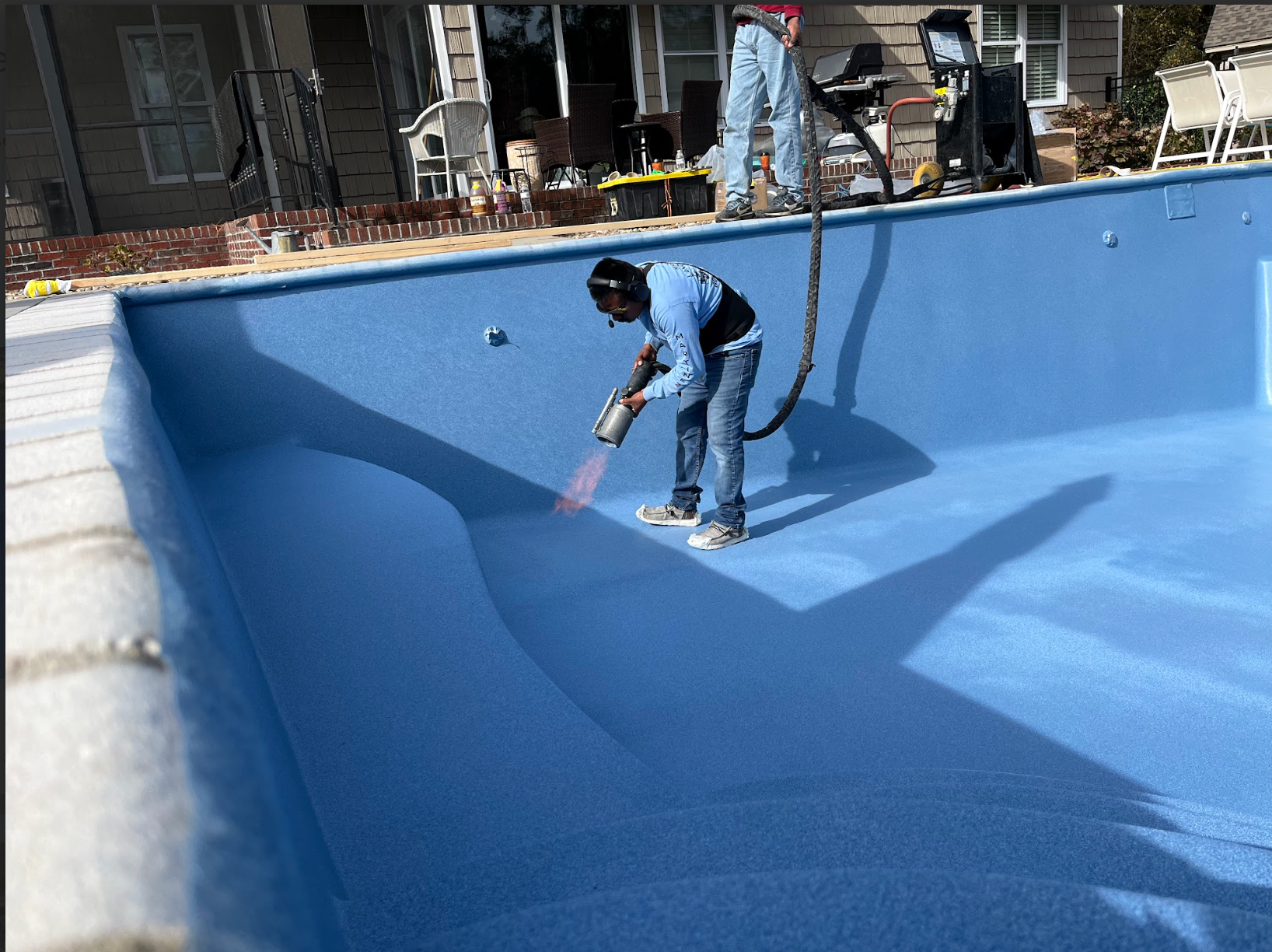 Man spraying blue coating inside an empty pool. Exterior setting with a house visible.