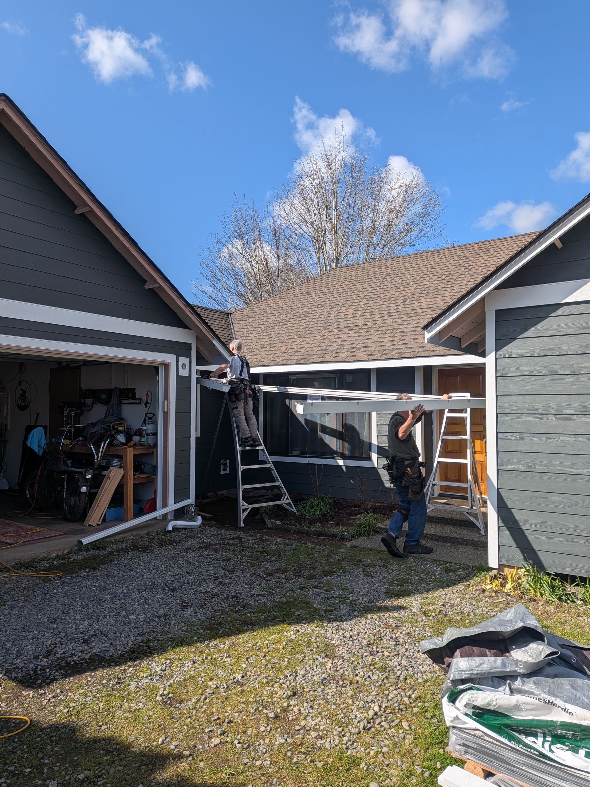 Person standing in a courtyard between garages, using a ladder to inspect roof damage under a blue sky.