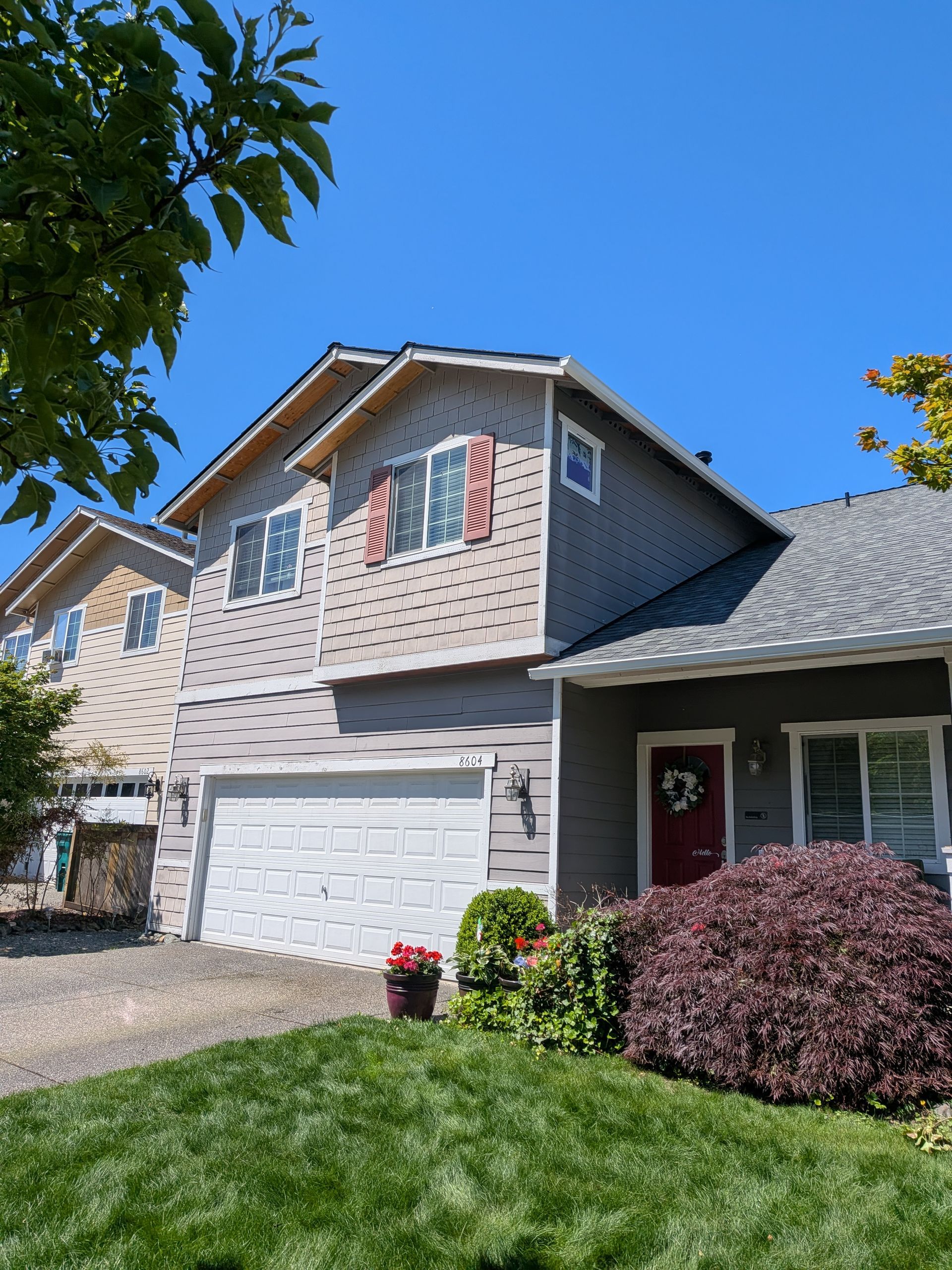 Suburban two-story house with gray siding, white garage door, red shutters, and a green lawn