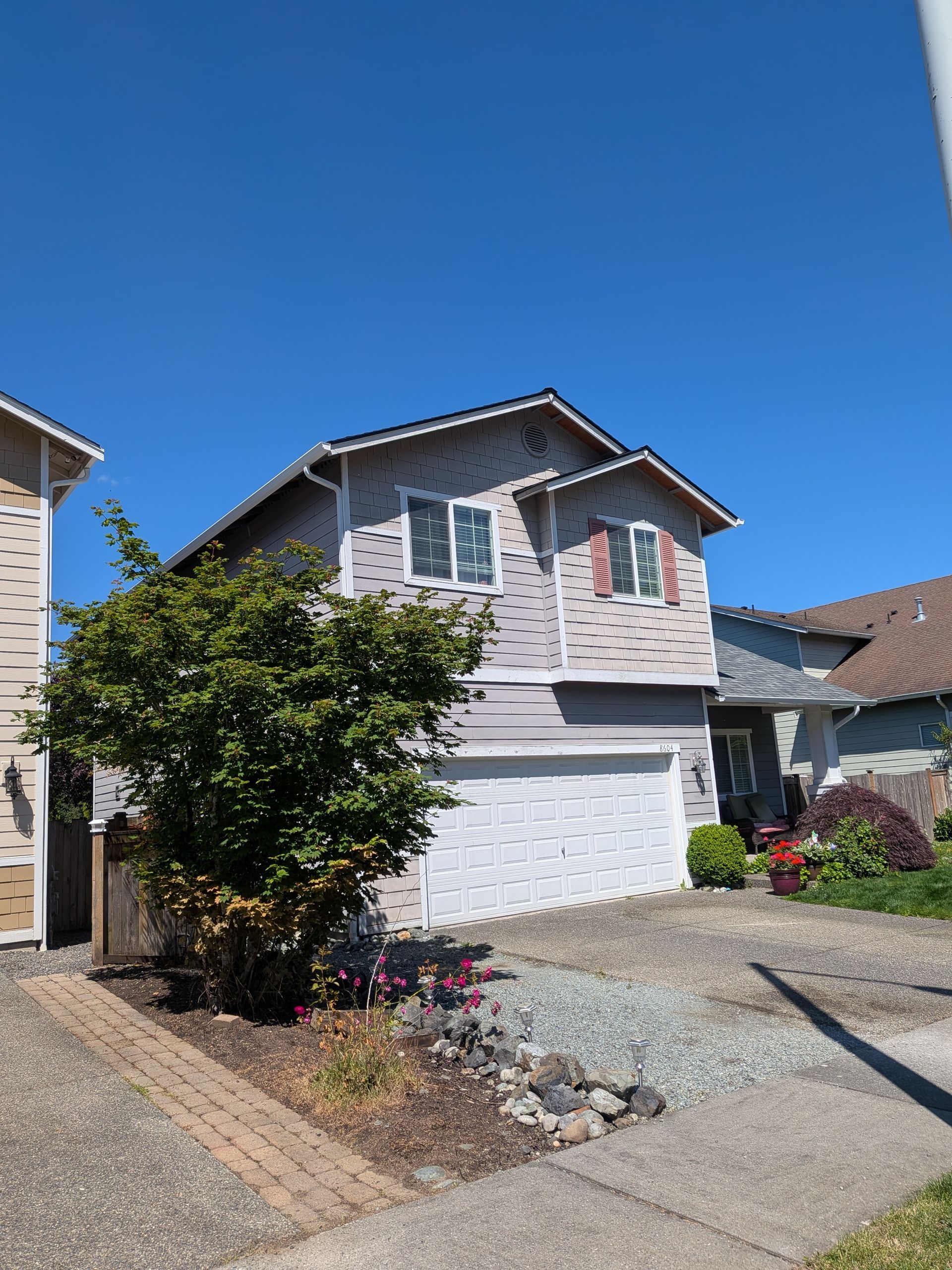 Suburban two-story house with white garage, gray siding, and small front garden under a clear blue sky
