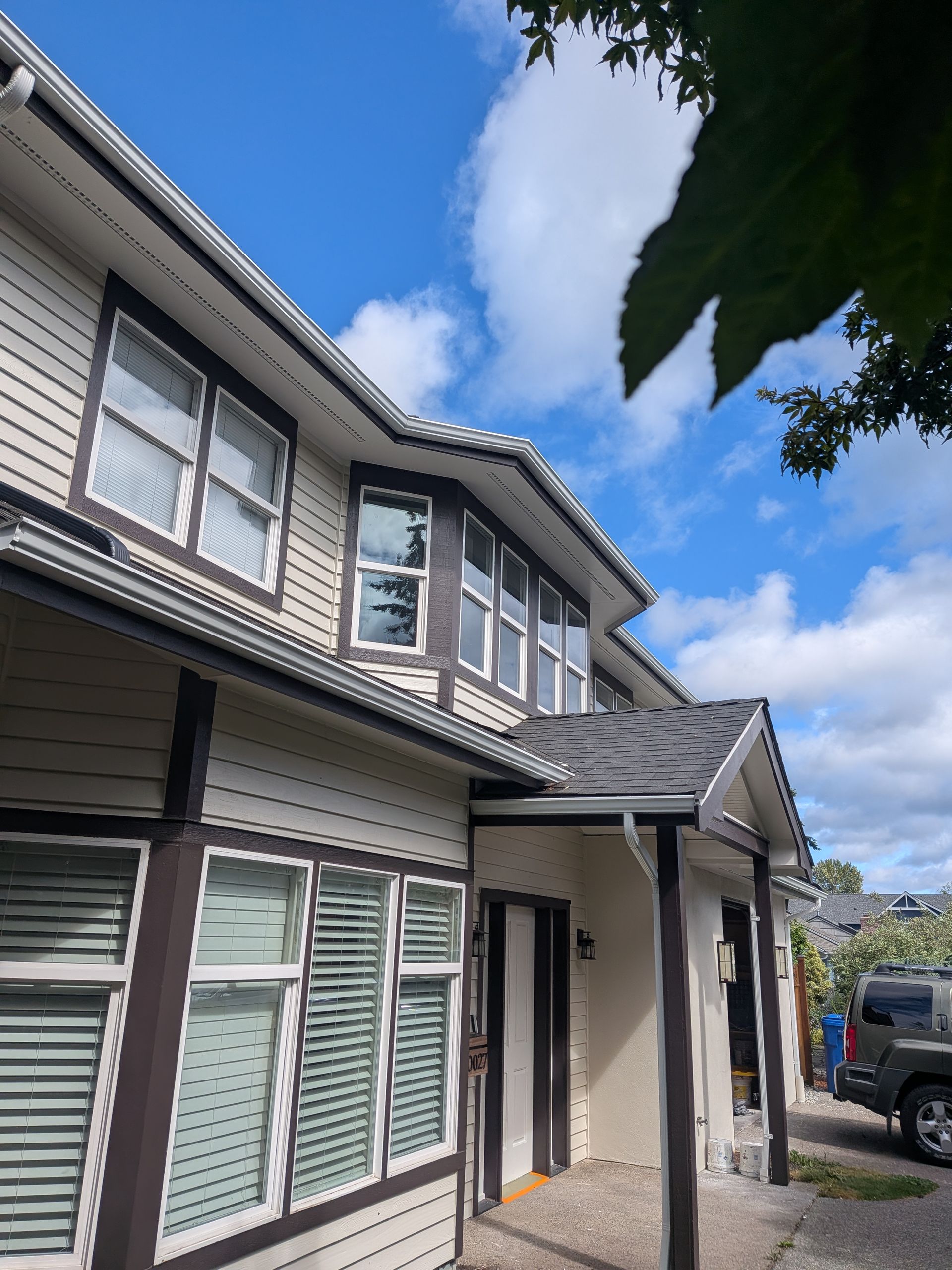 Two-story suburban house with gray siding, white trim, and a black car parked beside it under a blue sky.
