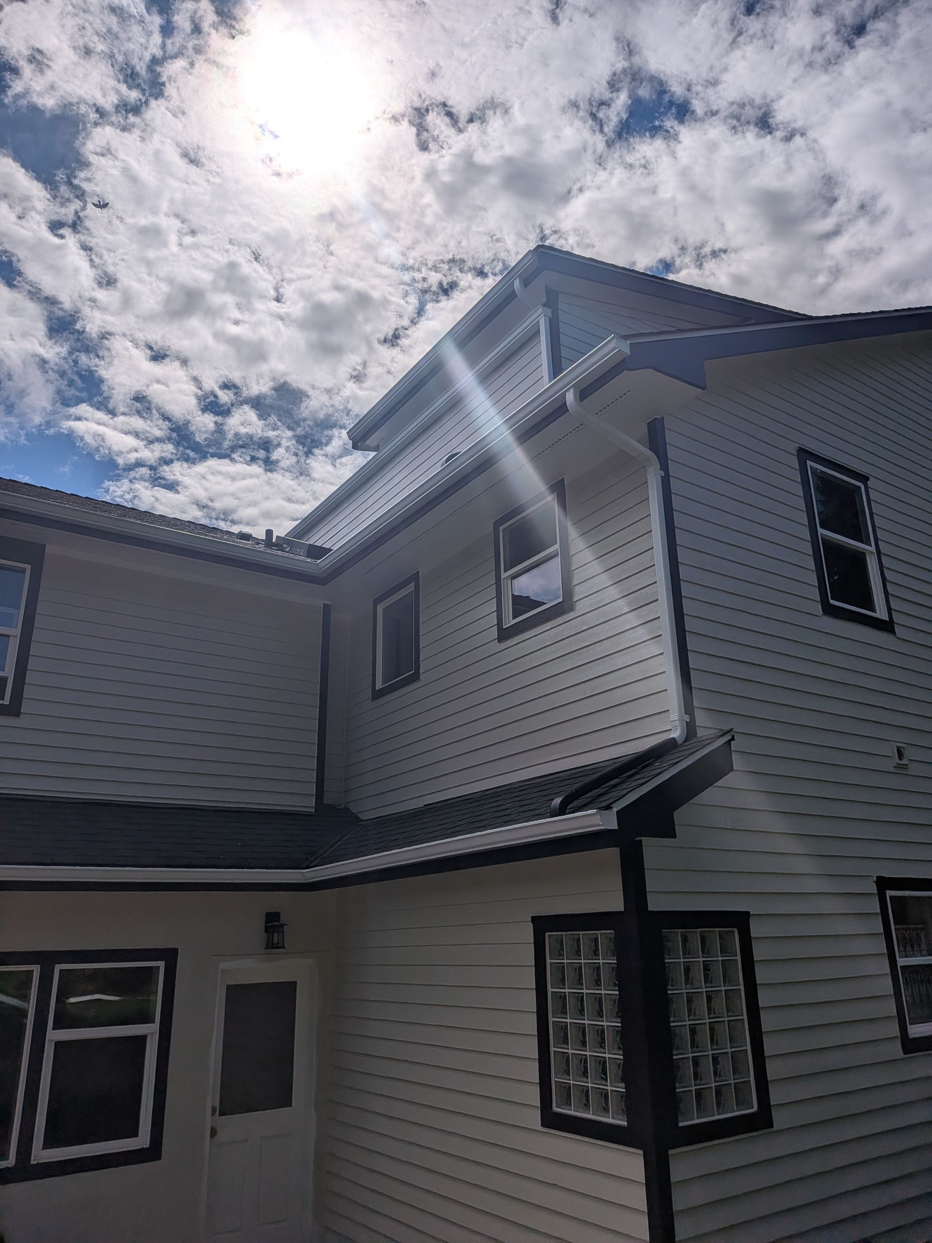 Two-story white house with black trim under a cloudy sky, sun flare near the roofline.