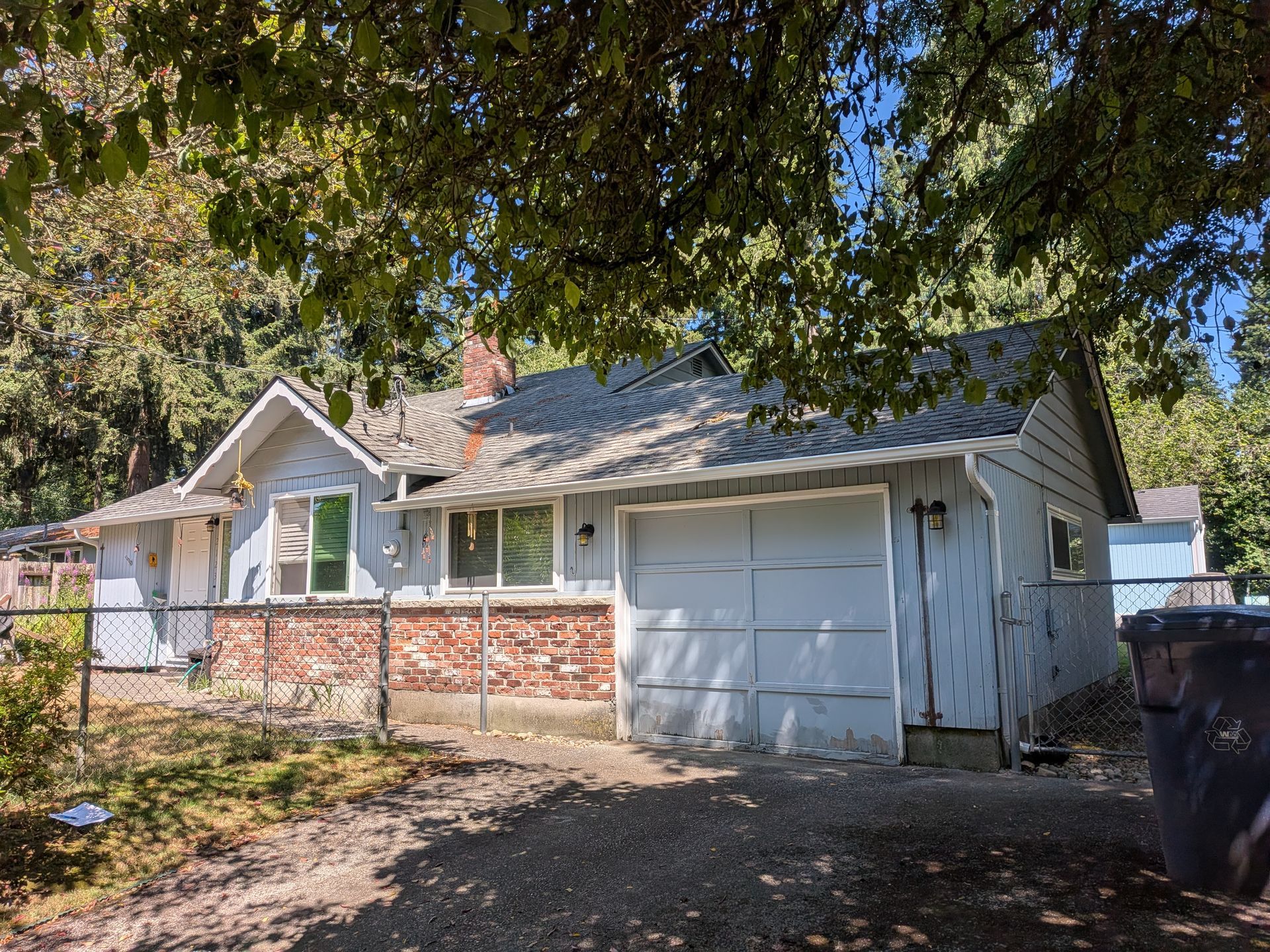 Small white house with a brick porch, garage door, and trees shading the front yard