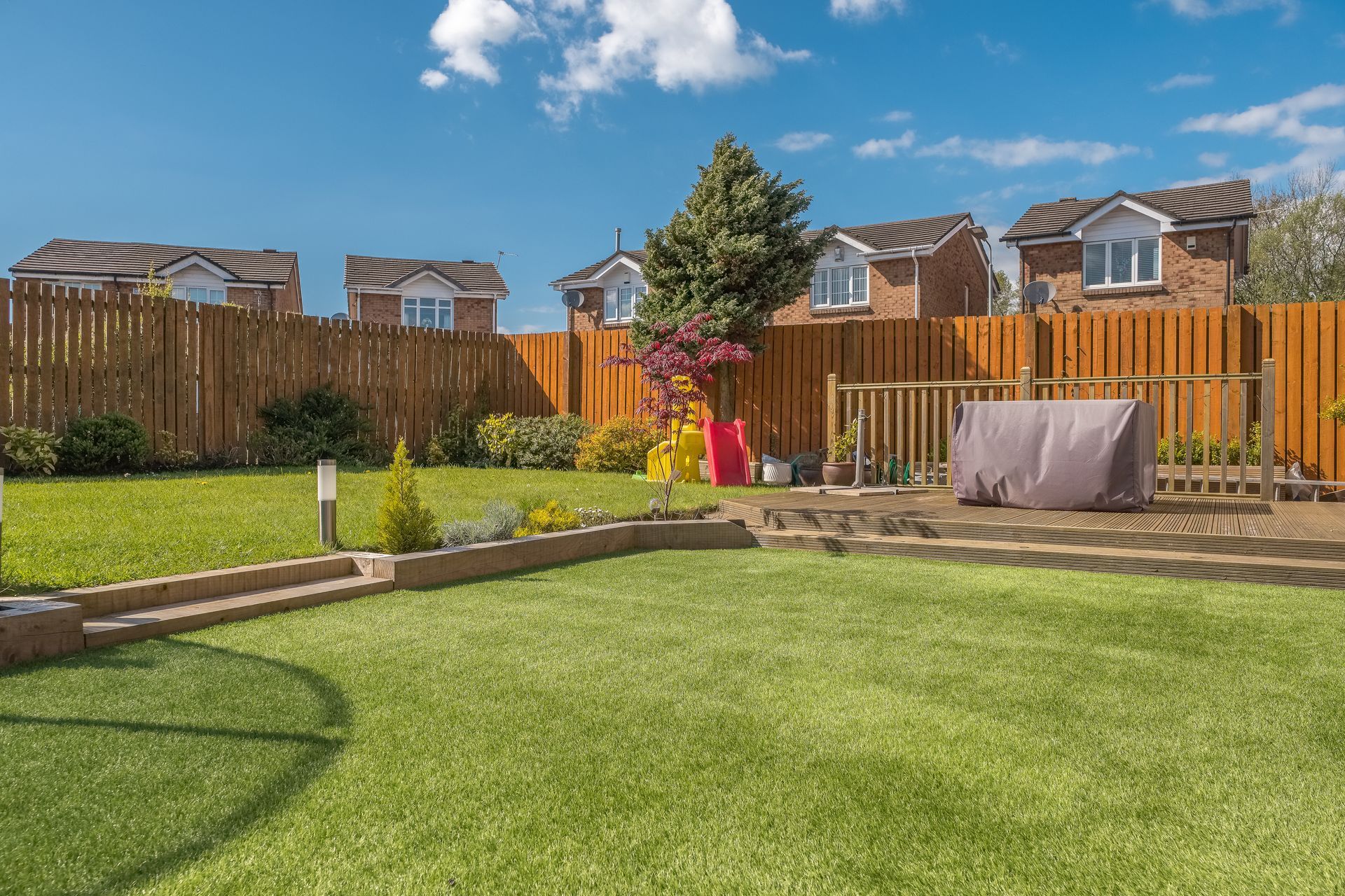Green backyard with lawn, wooden fence, houses, and bright blue sky.