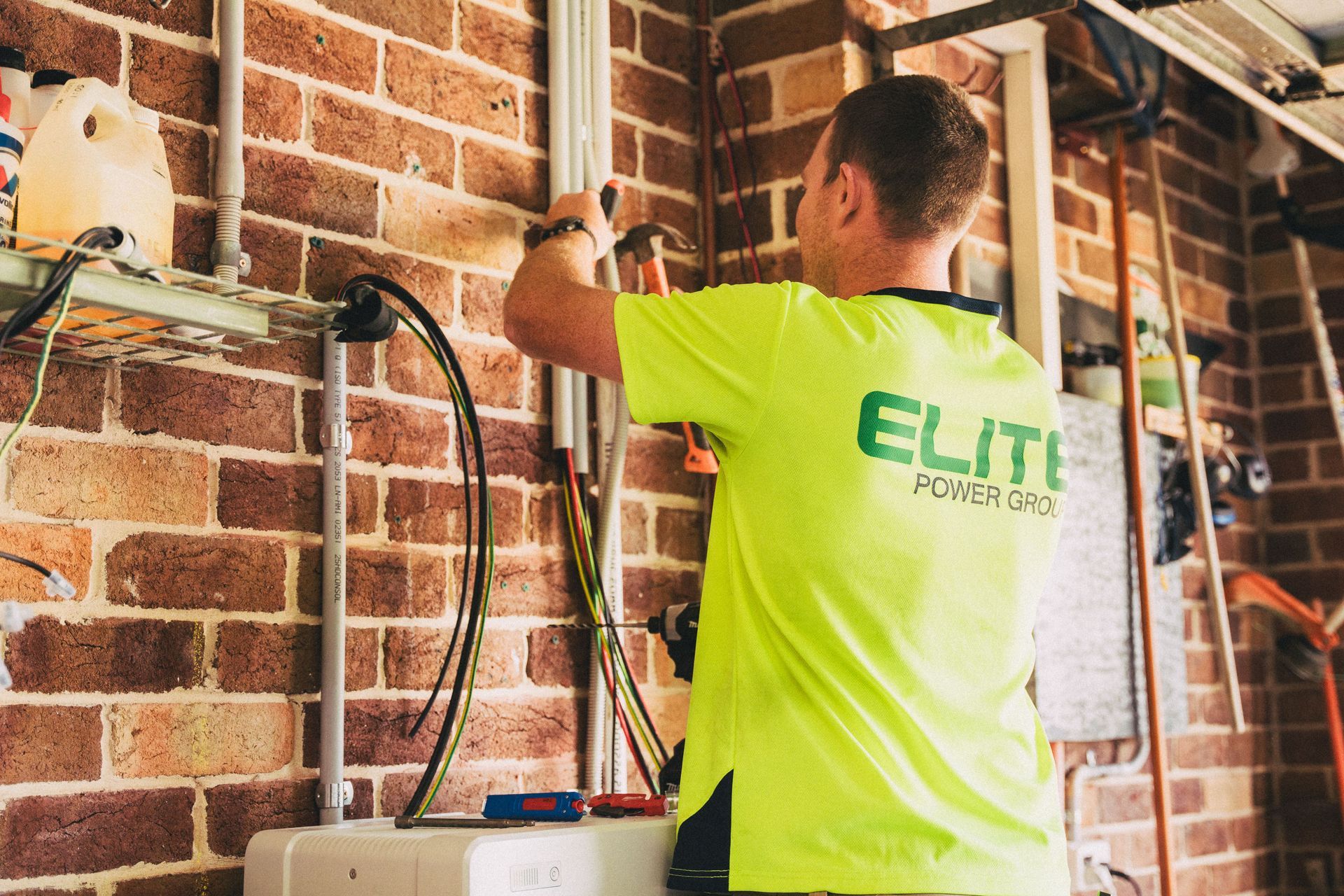 Electrician in neon green shirt wiring inside a brick-walled room with exposed studs