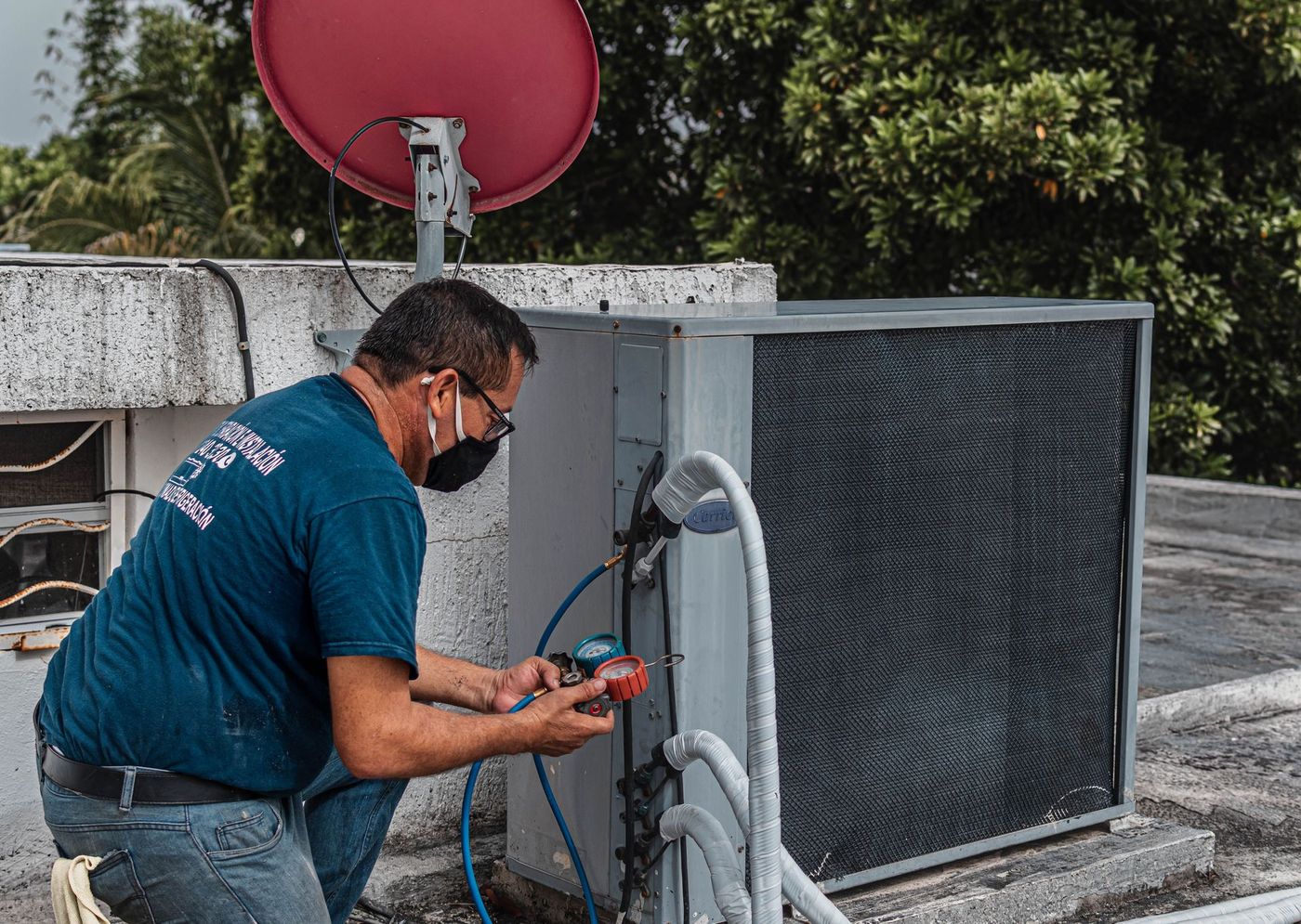Technician inspecting rooftop HVAC unit beside a red satellite dish and holding tools