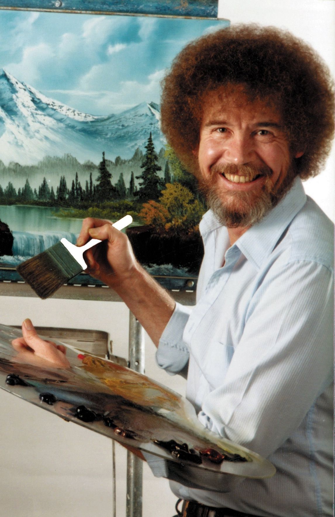 Bob ross is smiling while holding a brush in front of a painting