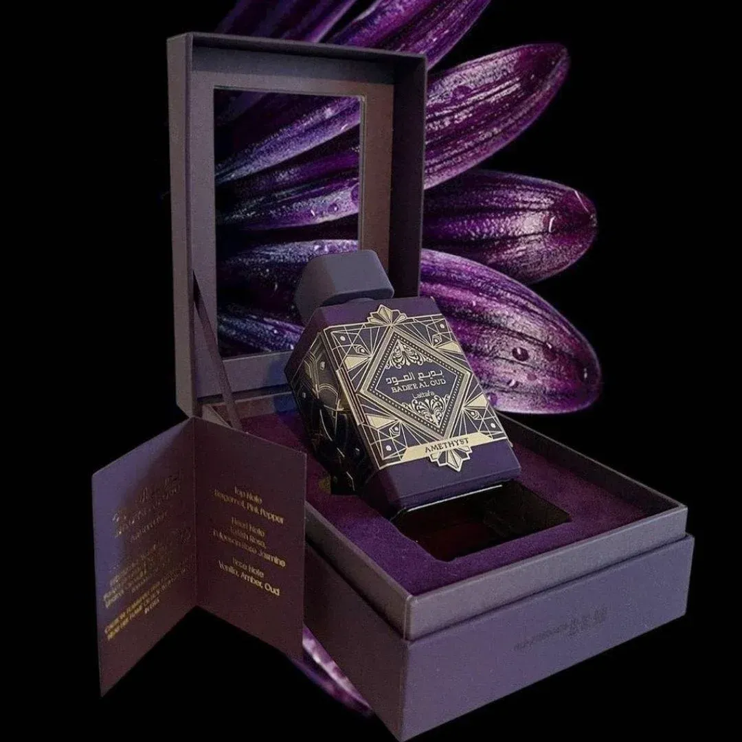 Frasco de perfume morado y caja a juego con diseño dorado de estilo árabe sobre fondo blanco.