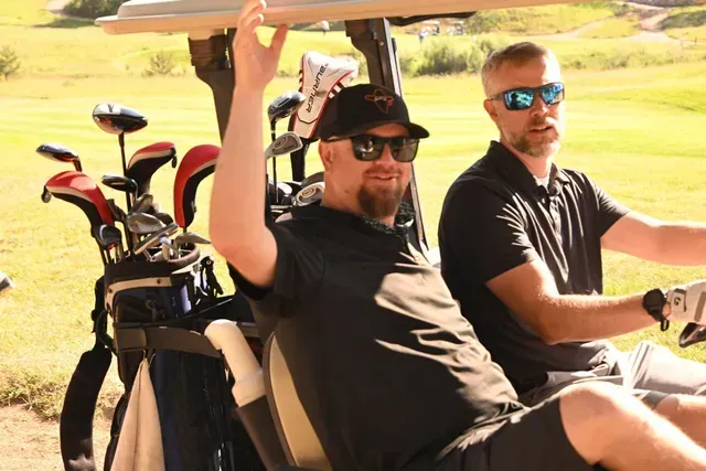 Two men in a golf cart on a course. One raises his arm, both wear sunglasses. Sunny day.
