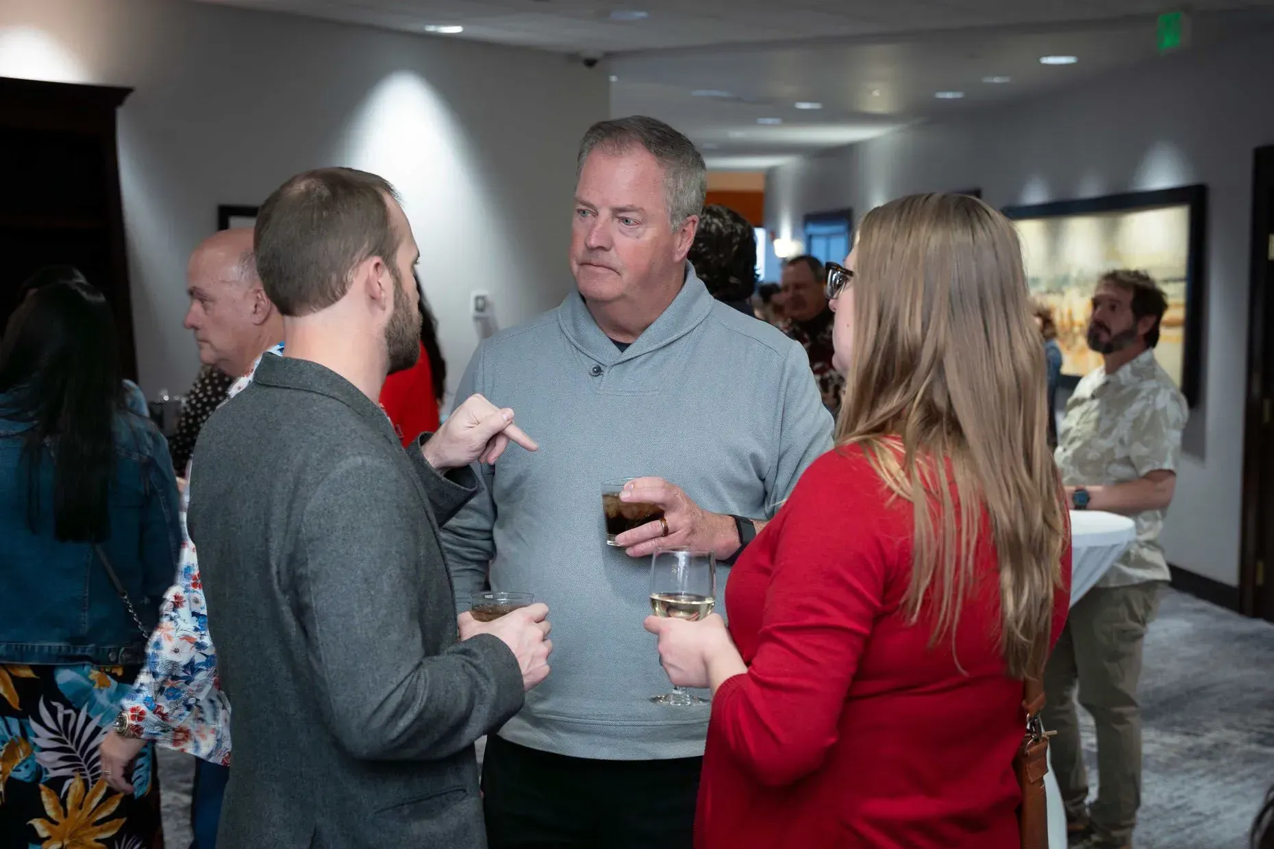 People in an event space chatting, drinks in hand. Man in gray sweater gestures, others listen.