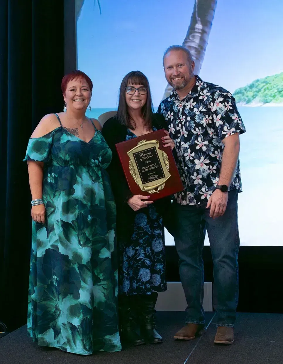Three people smiling, one holding a plaque on stage with a tropical backdrop.