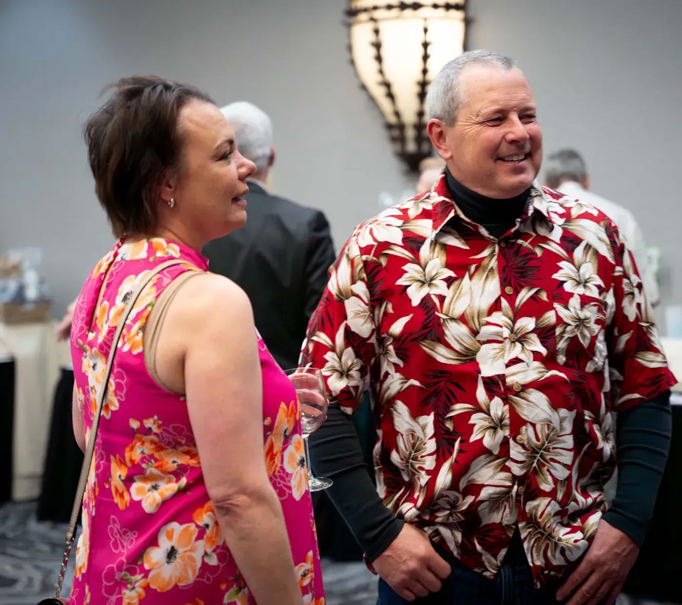 Woman in pink floral dress and man in red floral shirt smiling, chatting at an event.